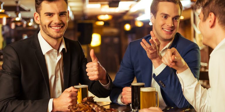 Three young businessmen in suits are smiling, talking and drinking beer while sitting in pub, dark haired man is showing Ok sign